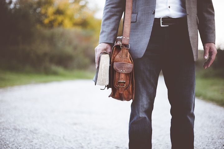 A torso down photo of a white man in business casual gray suiting carrying a thick book and his brown messenger bag as he walks down a verdant road. 