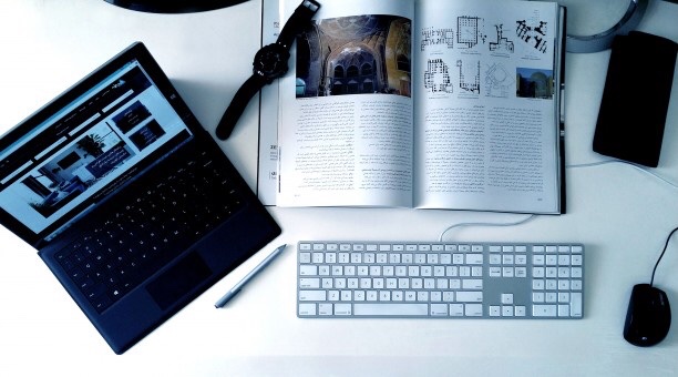 Laptop, magazine, keyboard, and notebook spread out across a workspace. 
